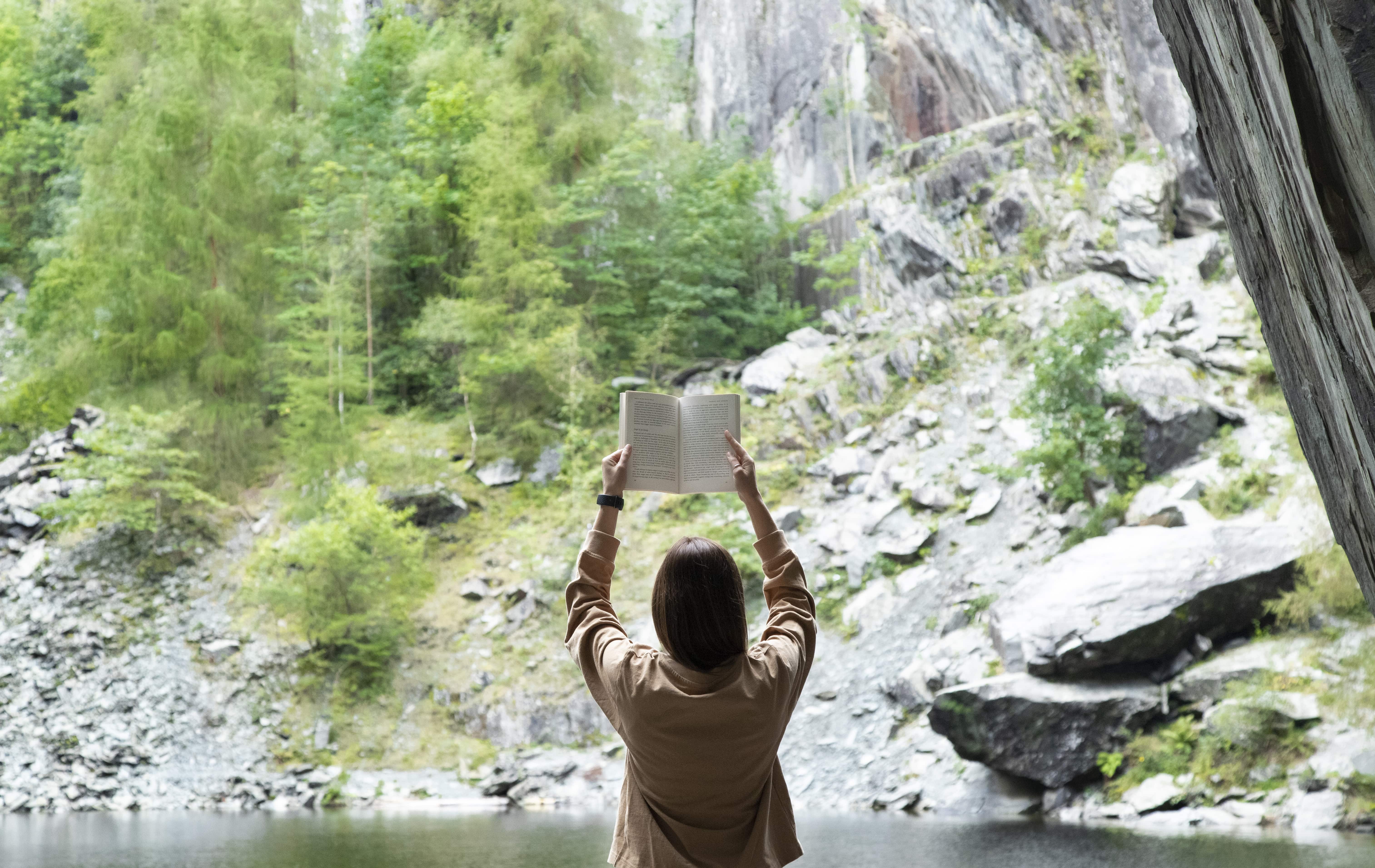 Liz holding a book above her head facing away, taken from inside a cave looking out to the hills